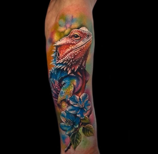 Colorful lizard tattoo on calf featuring a red-headed iguana with blue and gold body details perched above blue flowers with green leaves.
