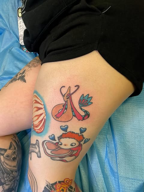 Colorful leg tattoo featuring whimsical creatures including a red-and-blue bird with extended wings, a character with orange hair, and other playful designs in coral, teal, and red tones.