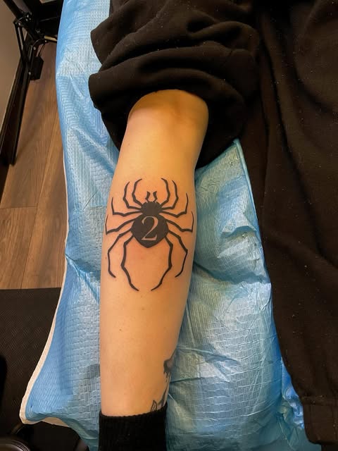 Black spider tattoo with number 2 in the center on a person's forearm.