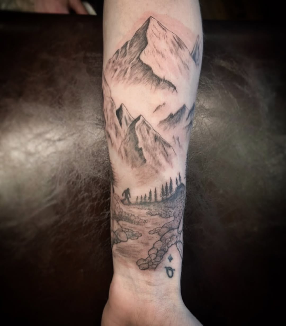 Mountain landscape tattoo on forearm featuring snow-capped peaks, forested valleys, and rocky terrain in black and grey shading.