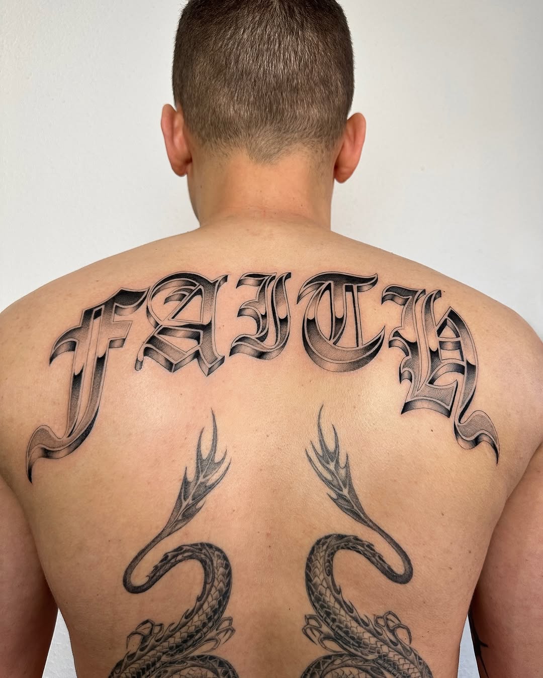 Back tattoo featuring ornate lettering at top, feathered designs, and detailed serpent motifs in black and grey shading.