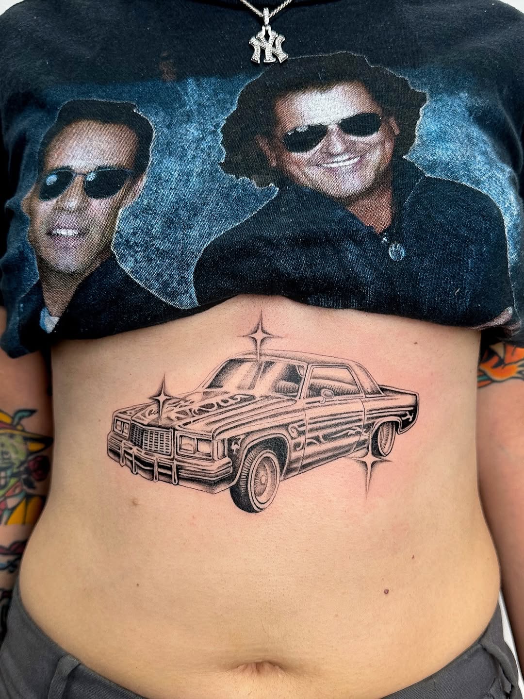 Black and grey lowrider car tattoo on chest with decorative cross above, smooth shaded realism style.