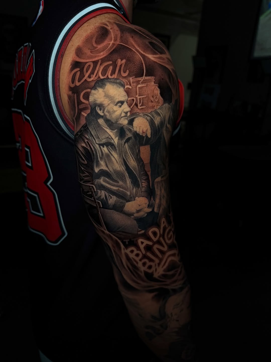 Portrait tattoo of a man in a leather jacket on upper arm, rendered in black and grey with soft shading and decorative text elements.