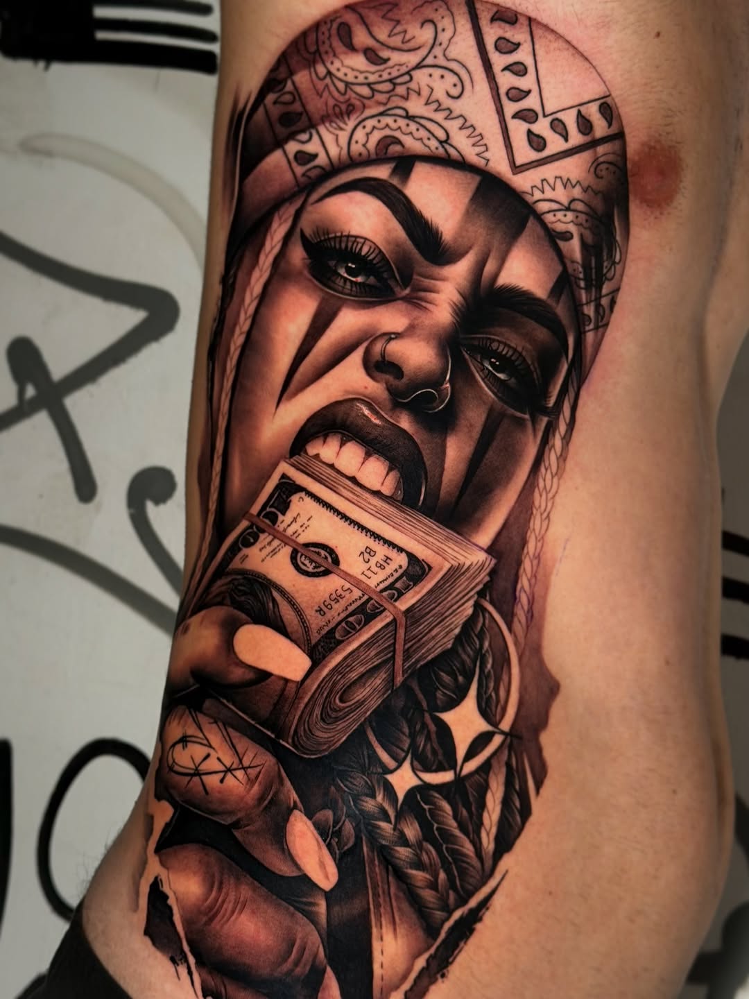 Black and grey portrait tattoo of a face wearing an ornate headdress, holding a stack of cash, on the arm.