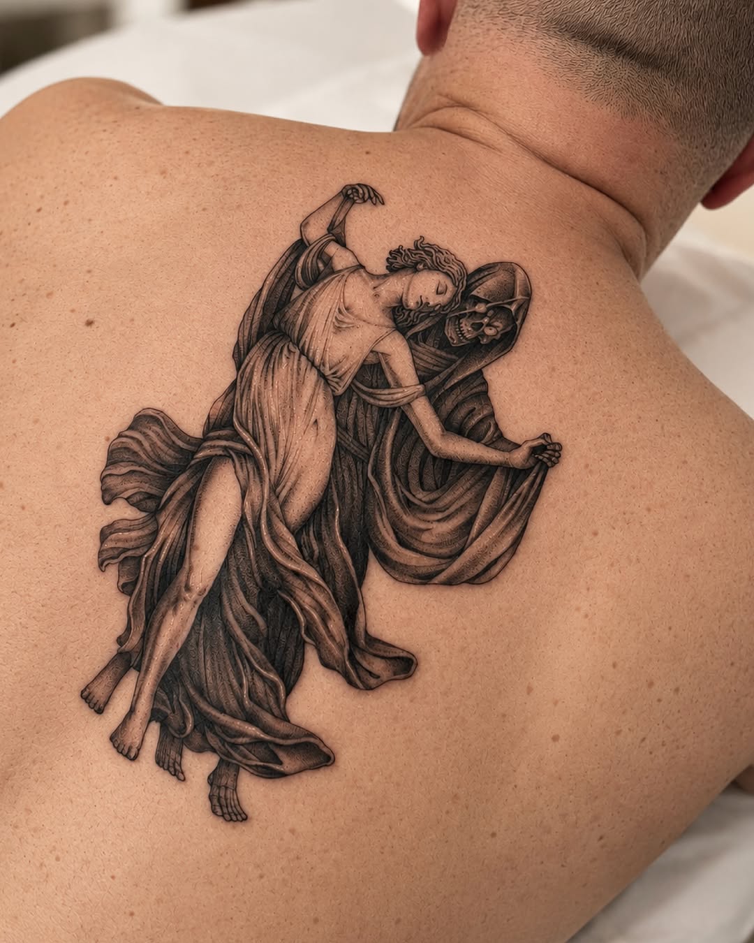 Black and grey winged angel figure in flowing robes with detailed shading, tattooed on shoulder blade area.
