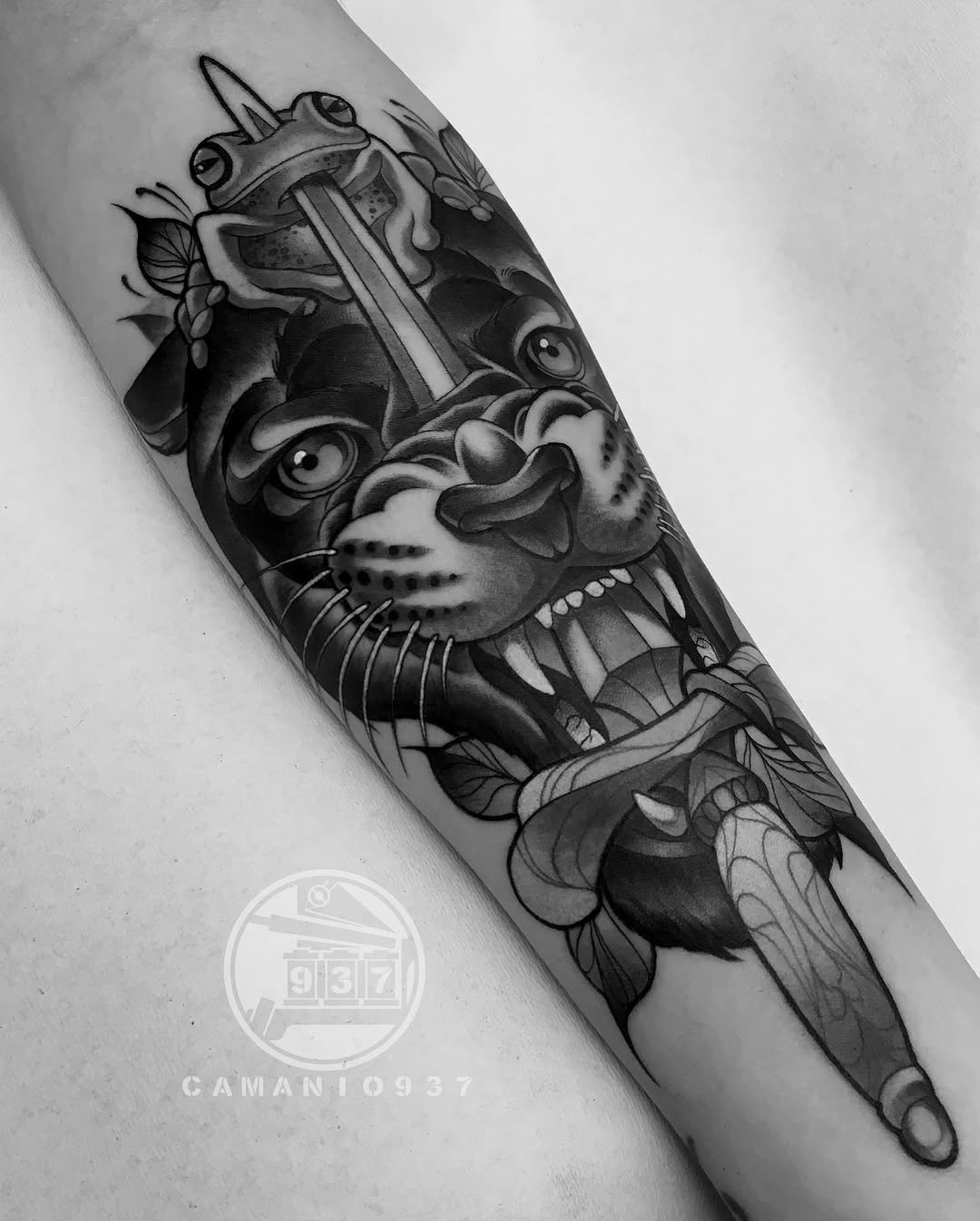 Black and grey forearm tattoo featuring a frog with ornamental mask and gun, surrounded by botanical elements and decorative details.
