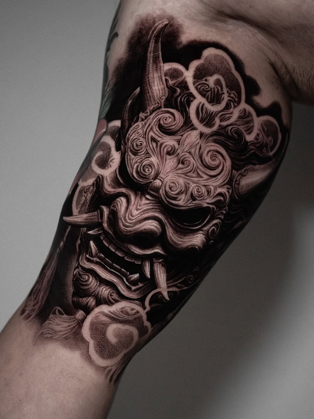Ornate demon mask tattoo with intricate swirling details on the upper arm, rendered in black and grey with smooth shading.