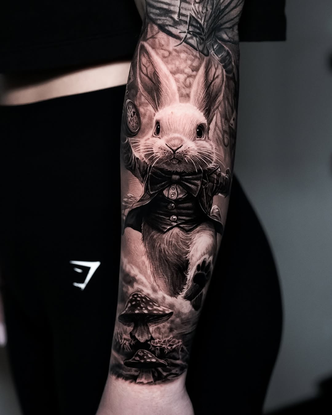 Anthropomorphic rabbit wearing a formal black bow tie and vest, tattooed on forearm with mushrooms below, rendered in black and grey with soft shading.