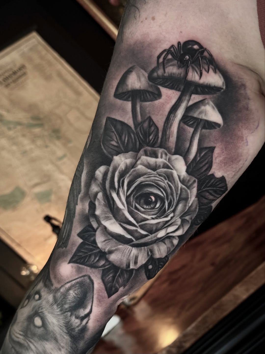Black and grey tattoo on forearm featuring a rose with an eye in its center, mushrooms above, and a spider, with soft shading throughout.