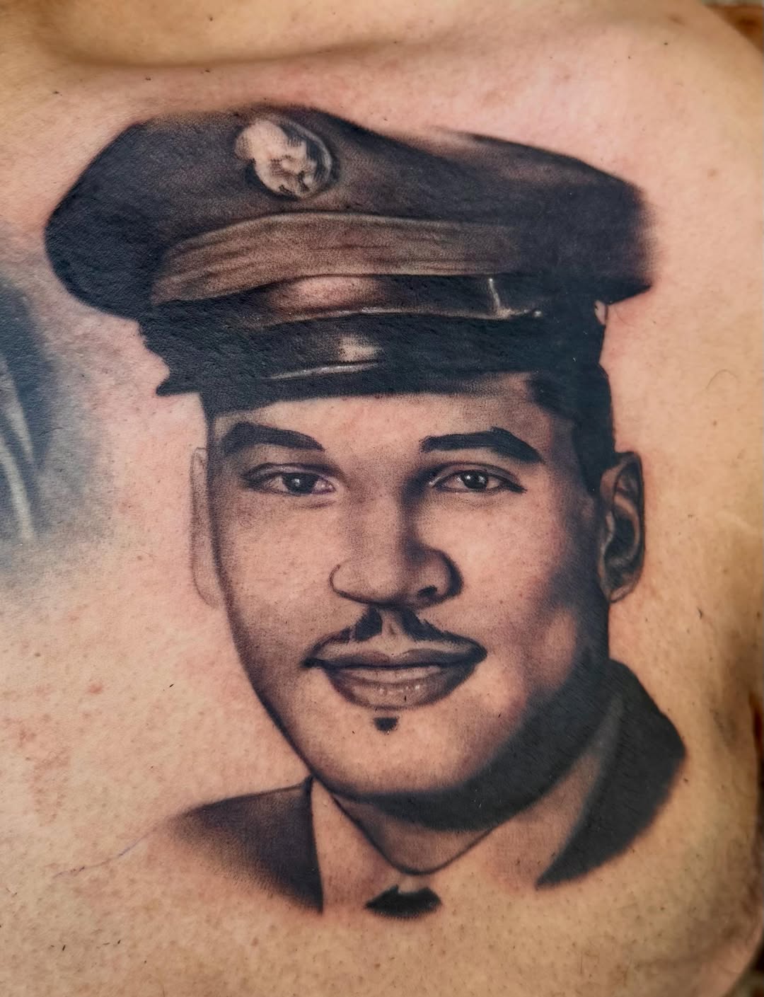 Black and grey portrait tattoo of a man wearing a military cap with insignia, featuring soft shading and realistic facial details on chest placement.
