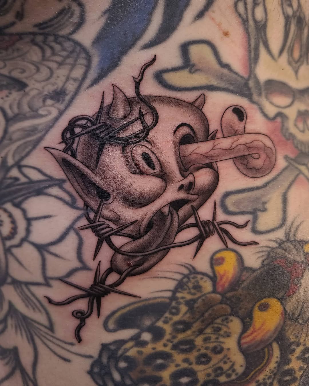 Black and grey tattoo of a demon pig head with horns and pointed ears on upper arm, surrounded by ornamental designs.