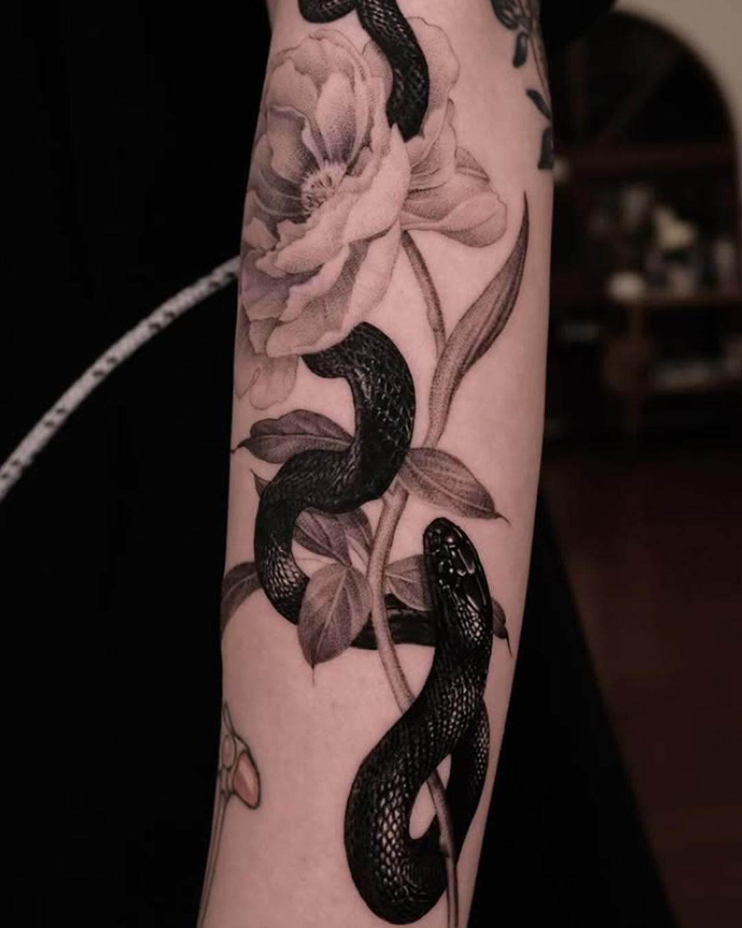 Black and grey tattoo on forearm featuring intertwining snakes and detailed flowers with soft shading and realistic botanical elements.
