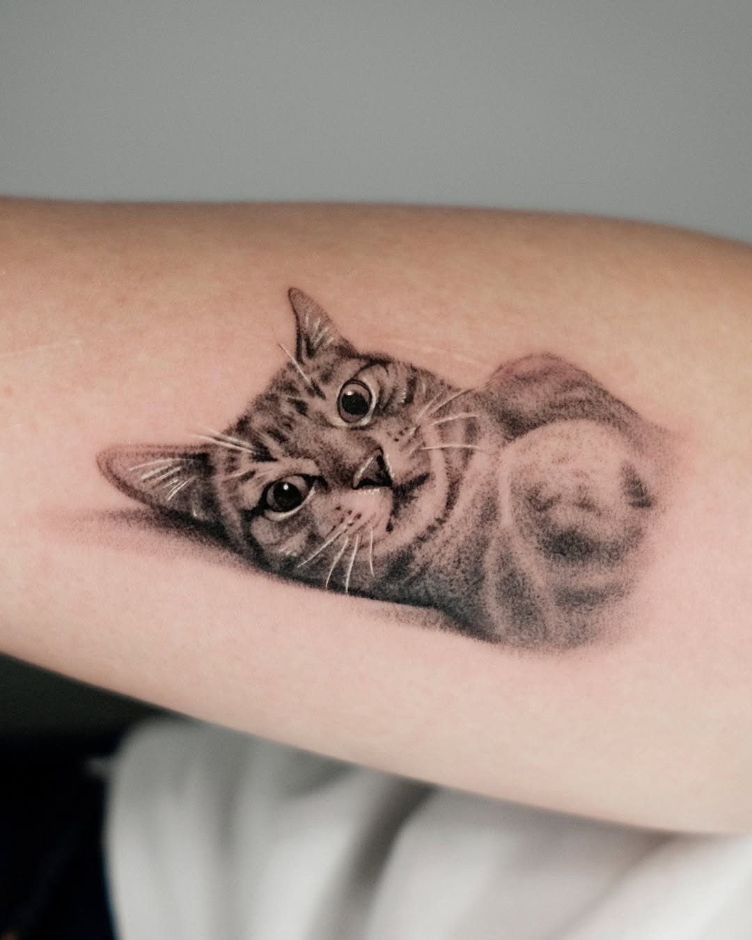 Tabby cat's face looking upward with detailed whiskers and expressive eyes, rendered in black and grey shading on forearm.