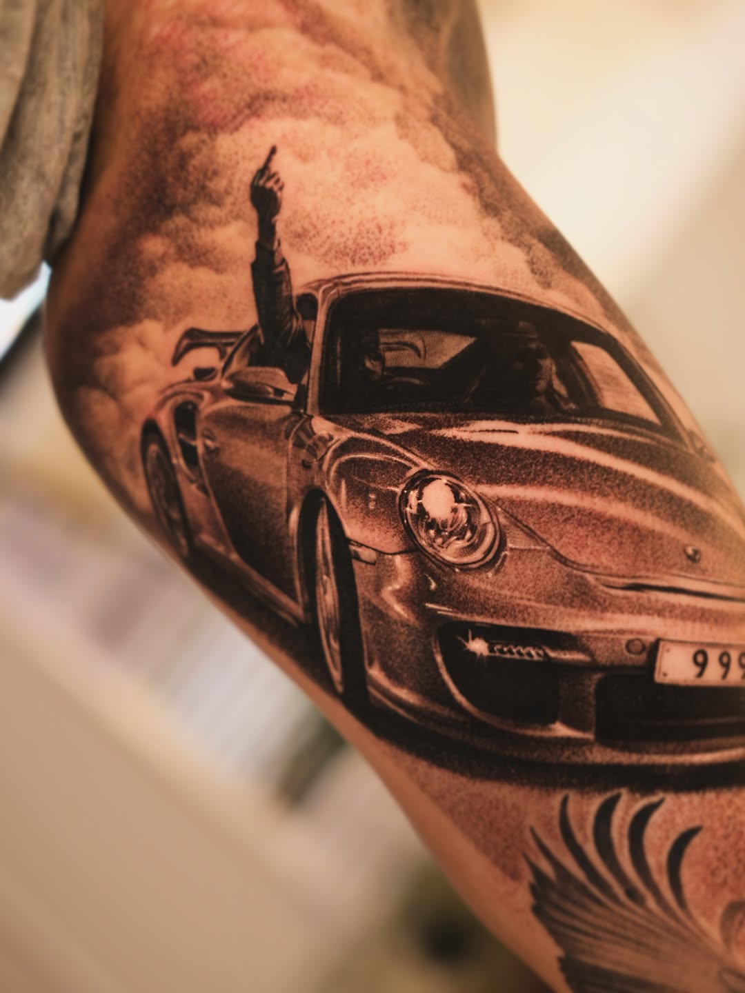 Realistic black and grey tattoo of a classic sports car with open door on forearm, showing detailed shading and gradients.