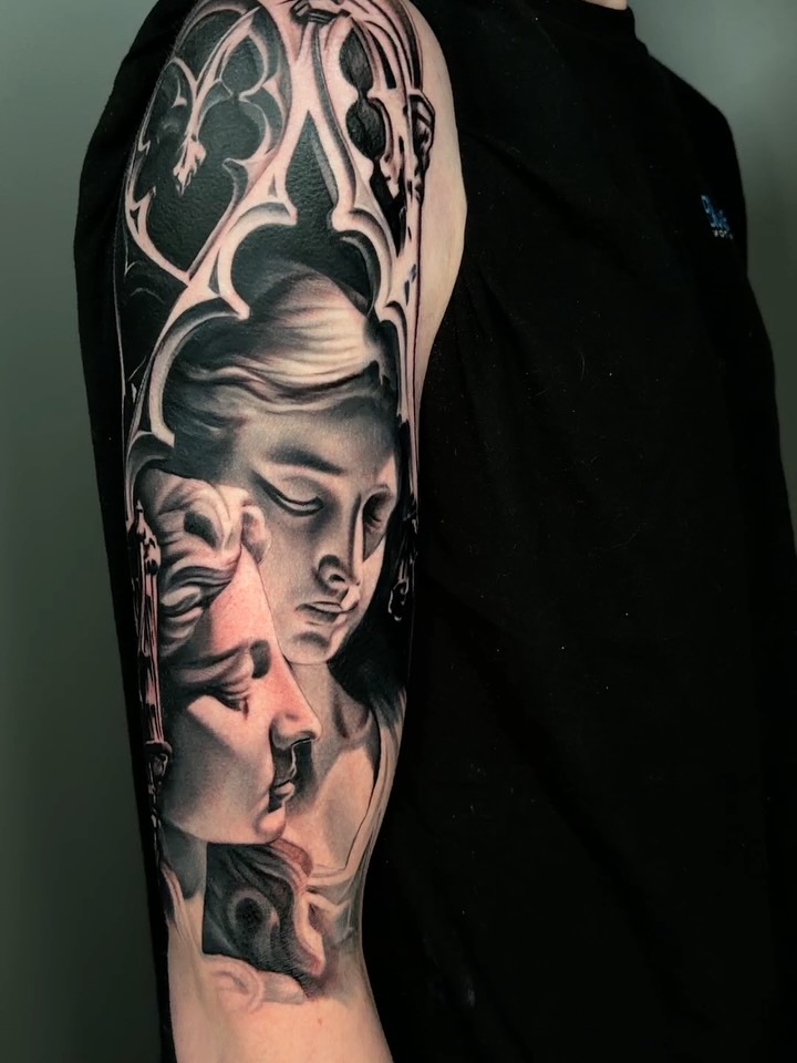 Multiple classical female faces with serene expressions and ornate decorative elements tattooed on upper arm in black and grey with soft shading.