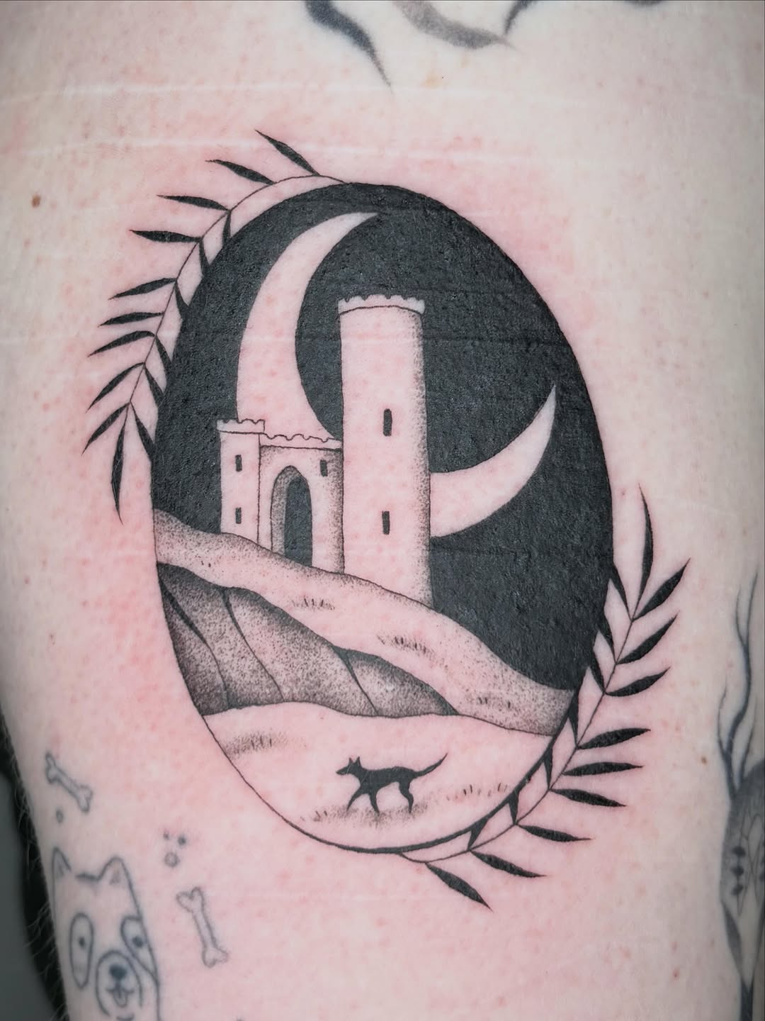 Black and grey tattoo of a crescent moon containing a fortress tower with a small silhouette of a dog, surrounded by botanical leaf elements.