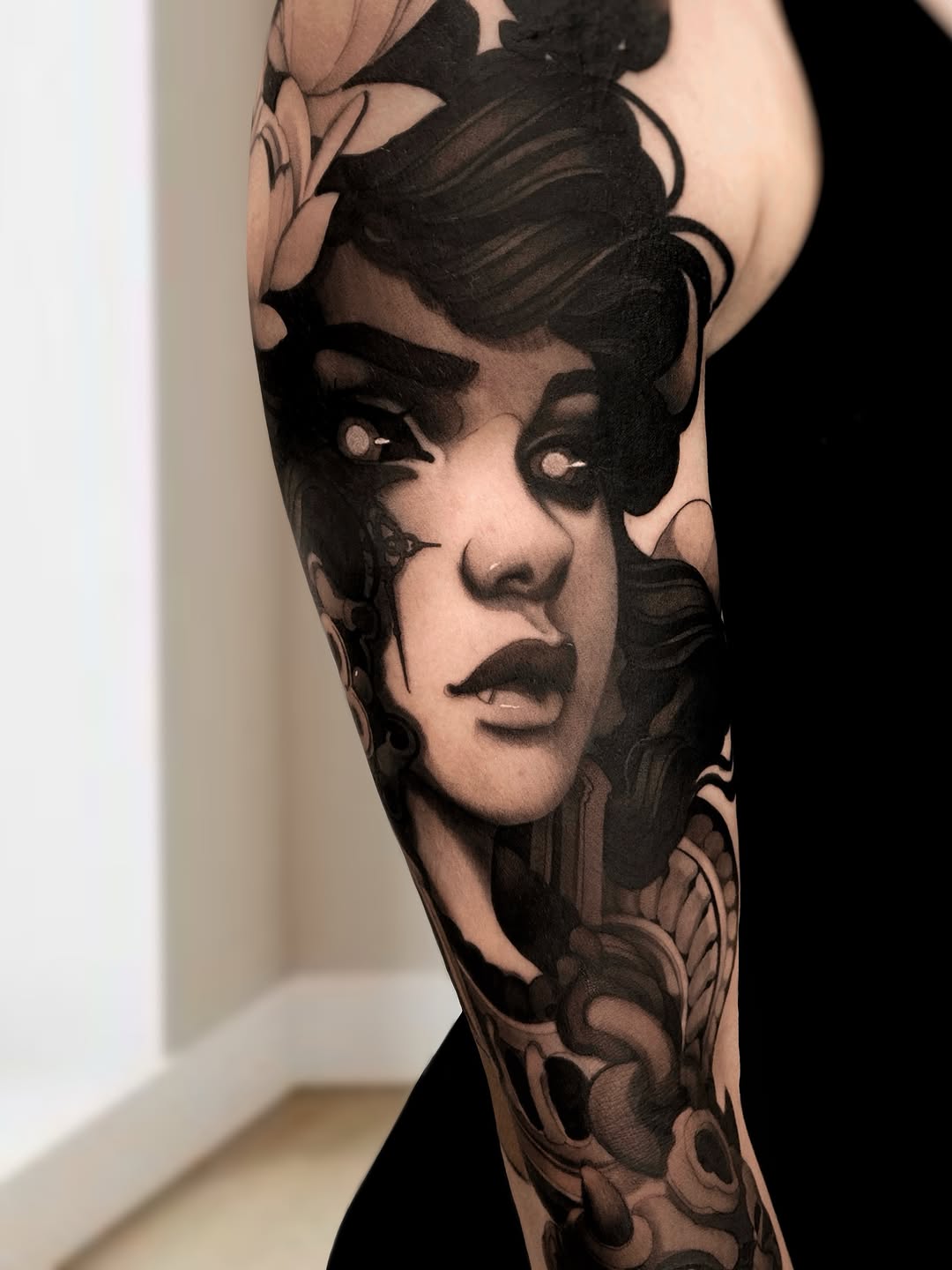 Black and grey portrait tattoo of a woman's face with dramatic shading on an upper arm, surrounded by decorative floral elements.