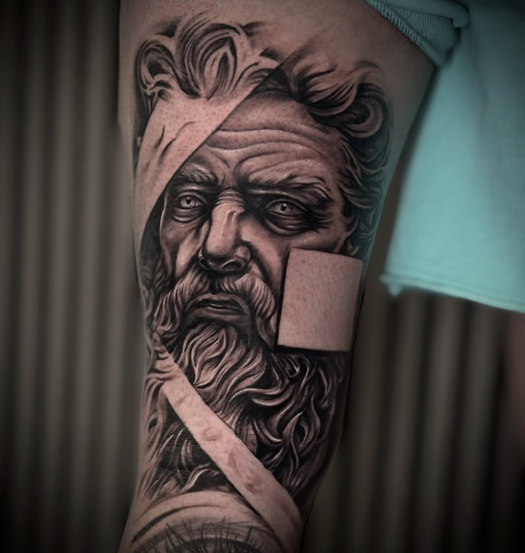 Detailed black and grey portrait of a bearded man with wavy hair on the upper arm, featuring smooth shading and realistic facial features.