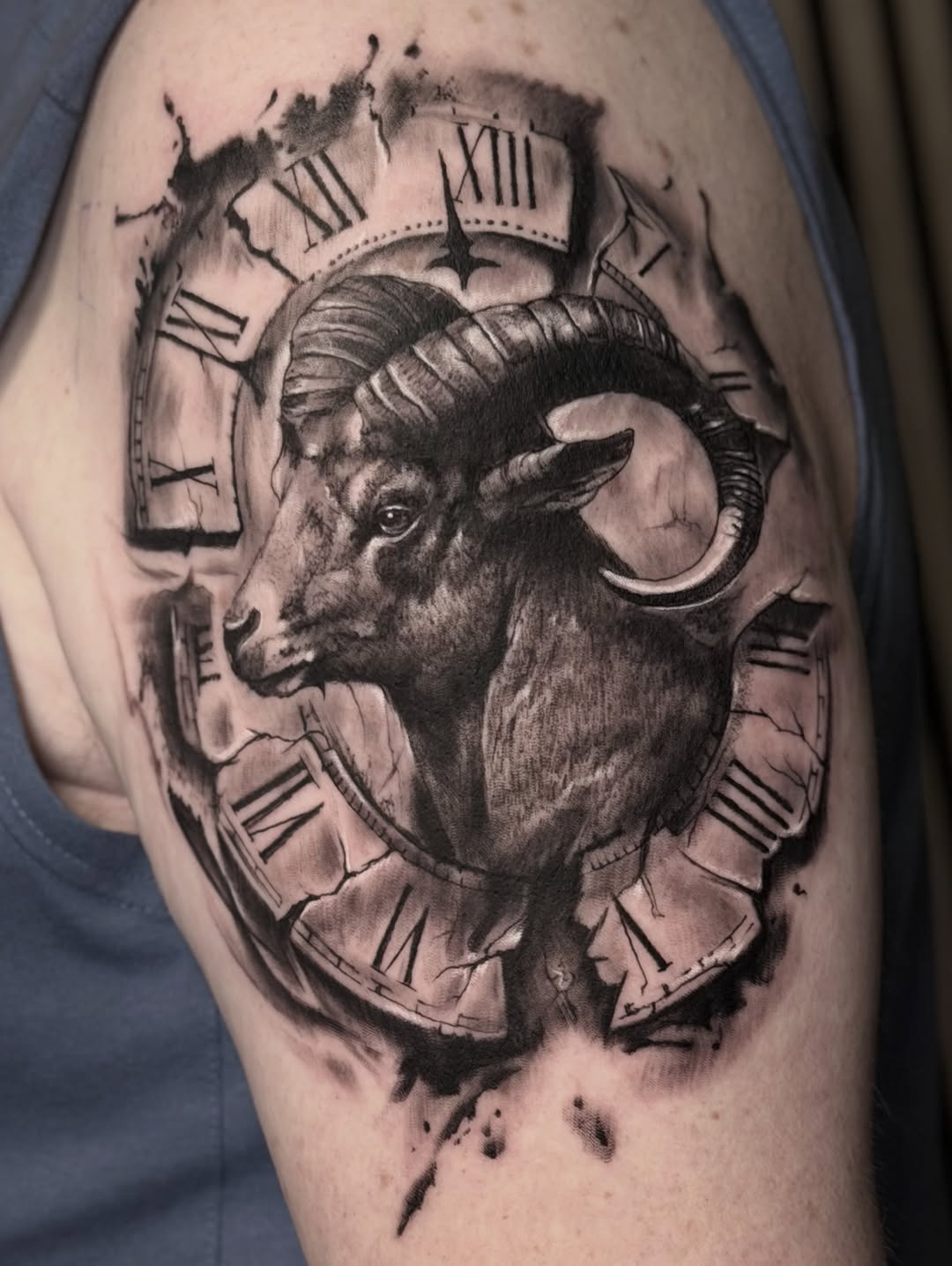 Black and grey ram's head tattoo with large curved horns surrounded by Roman numeral clock faces on upper arm.