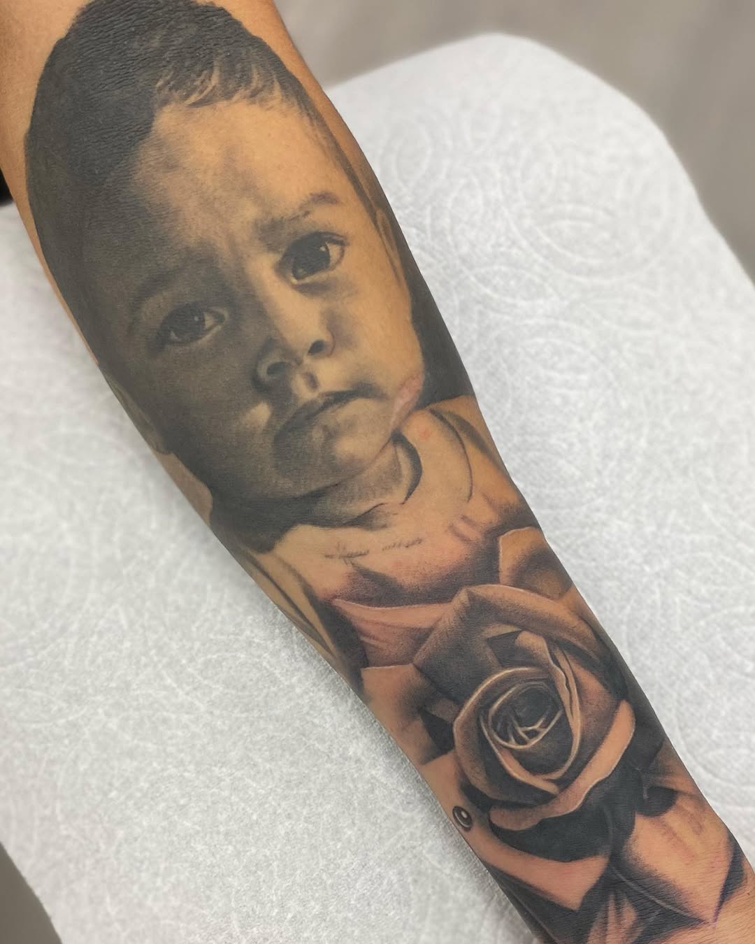 Portrait of a young child with contemplative expression on forearm, accompanied by a detailed rose below in black and grey shading.