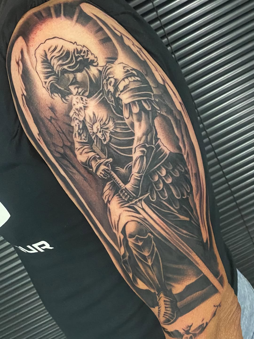 Saint Michael the Archangel in armor defeating a demon, black and grey realism tattoo on upper arm with detailed shading and religious iconography.