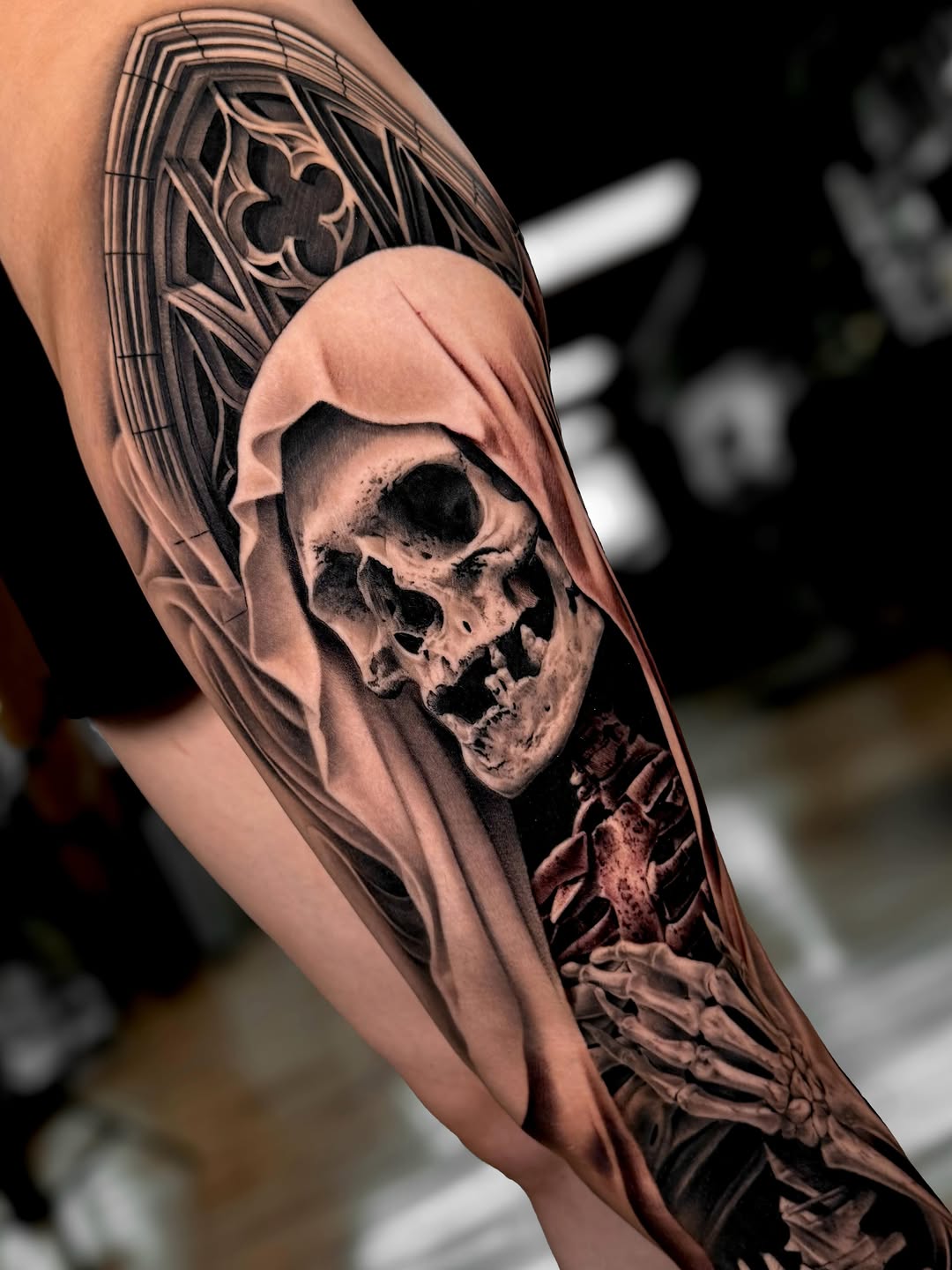 Grim reaper skull in hooded robe with ornate geometric patterns above, tattooed on forearm in black and grey with realistic shading.