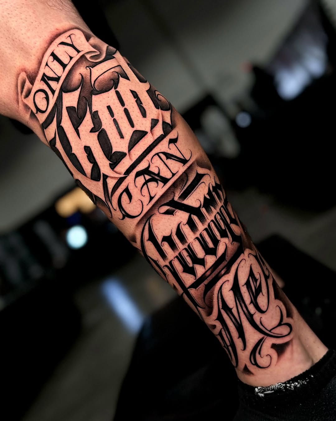 Black and grey sleeve tattoo featuring skulls with ornamental lettering and decorative flourishes on the forearm and bicep.