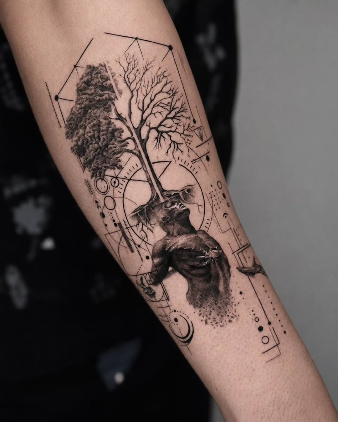 Forearm tattoo featuring a tree with full foliage on one side and bare branches on the other, with geometric shapes and a wolf head in black and grey shading.