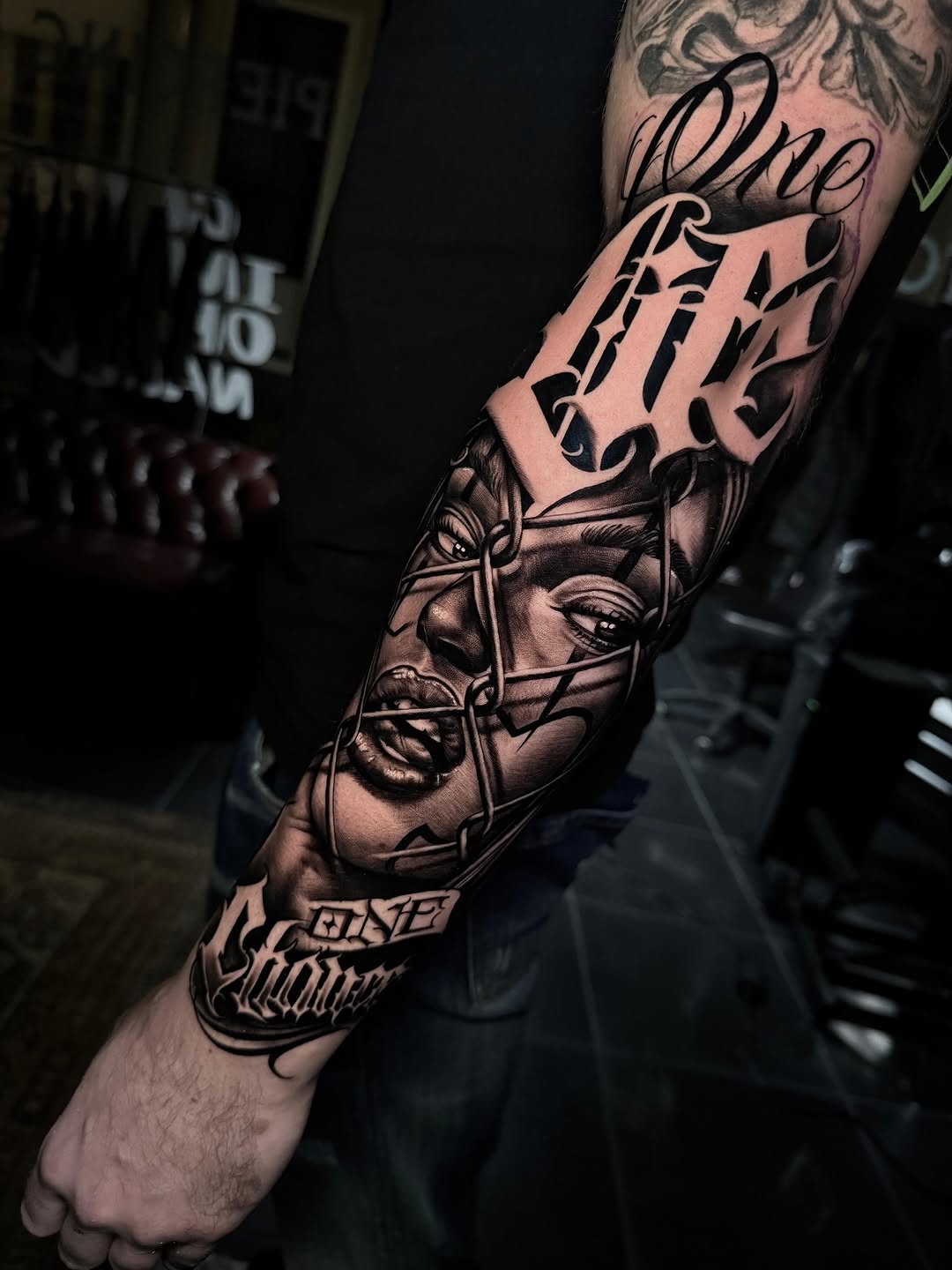 Black and grey sleeve tattoo featuring stylized faces with ornamental lettering and decorative elements on the forearm and upper arm.