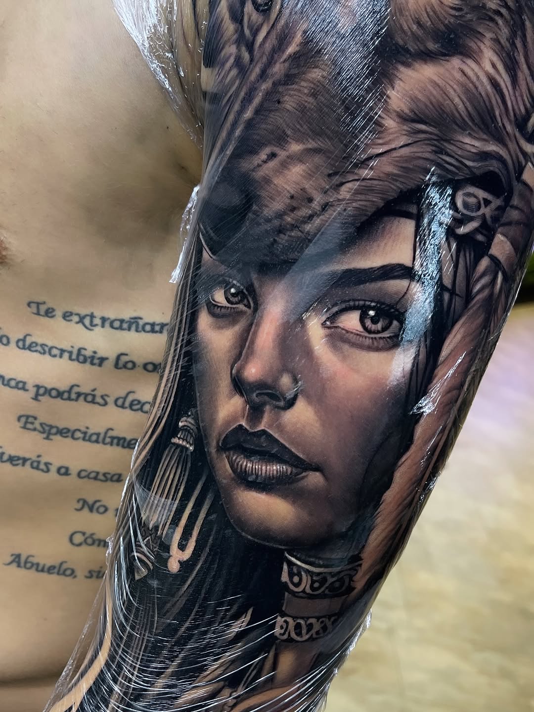 Black and grey portrait tattoo of a woman's face with detailed shading, long hair with feather elements, and decorative neck ornaments on the forearm.