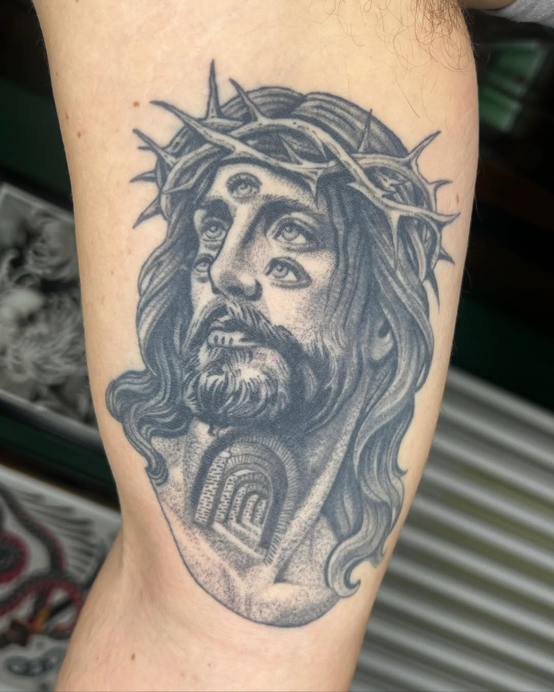 Black and grey portrait tattoo of Jesus Christ with crown of thorns, long hair and beard, on upper arm with architectural detail on chest.