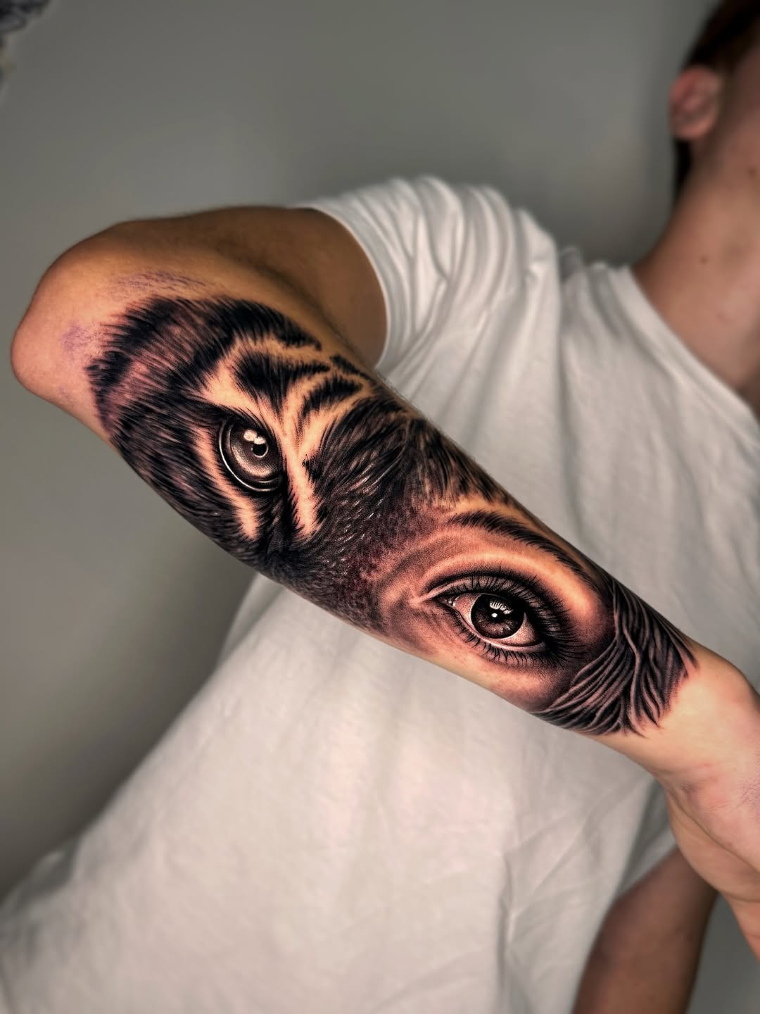 An owl's face and human eye rendered in black and grey with soft shading on the forearm, showcasing realistic detail and dimension.