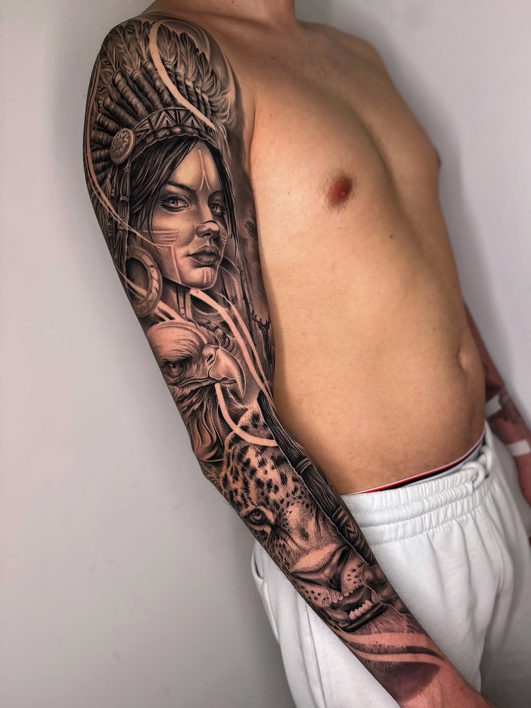 Full arm sleeve tattoo featuring a woman with elaborate feathered headdress and a leopard below in black and grey shading.