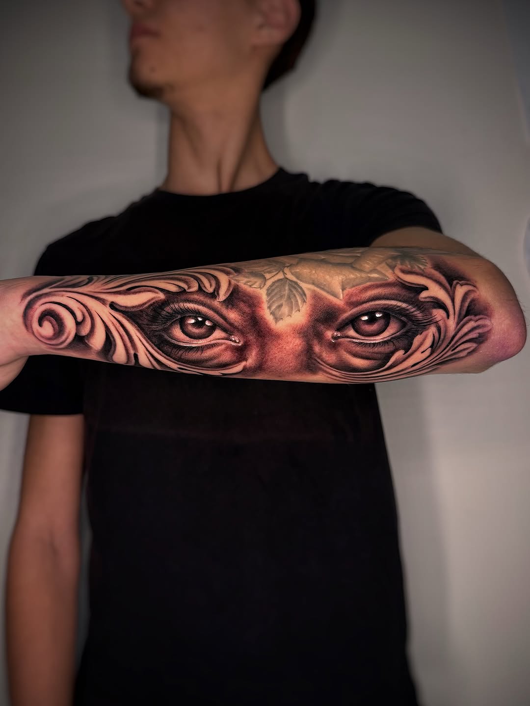 Realistic portrait tattoo of intense eyes with ornate flourishes on forearm, rendered in black and grey with smooth shading.