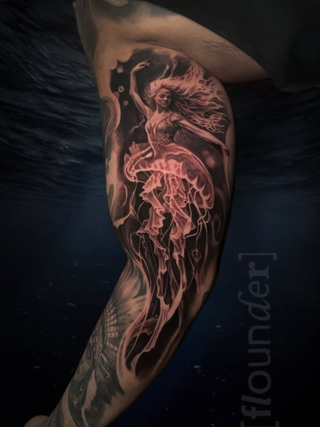 A mermaid with flowing hair and detailed jellyfish tail in black and grey shading on a leg.