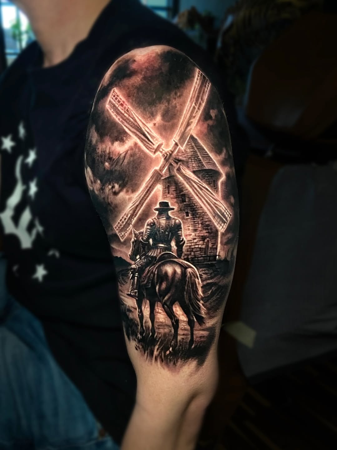 Upper arm tattoo depicts a knight on horseback facing a windmill under a glowing sky, rendered in black and grey shading.