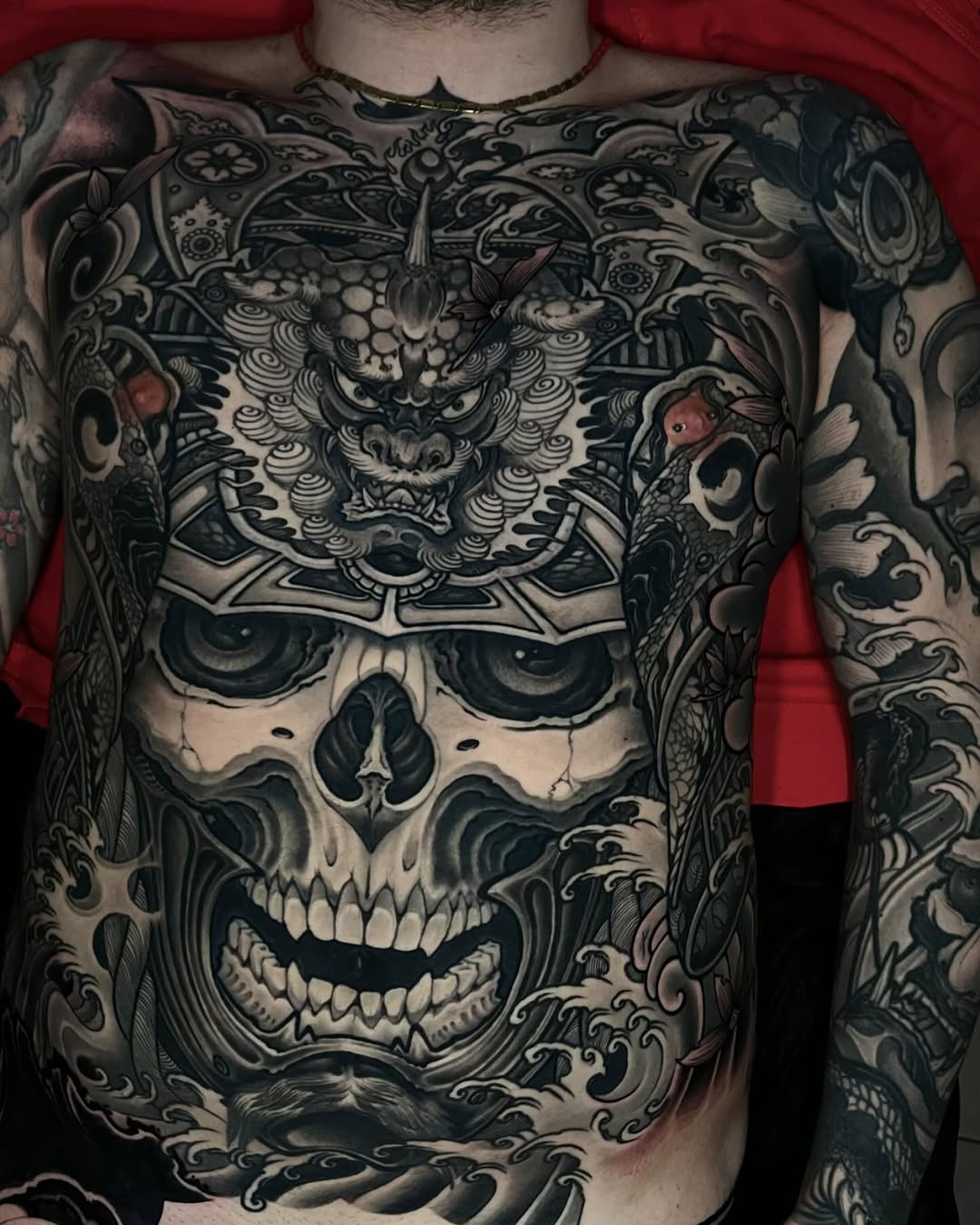 Ornate chest and sleeve tattoo featuring a demon mask above a skull with elaborate baroque scrollwork in black and grey.