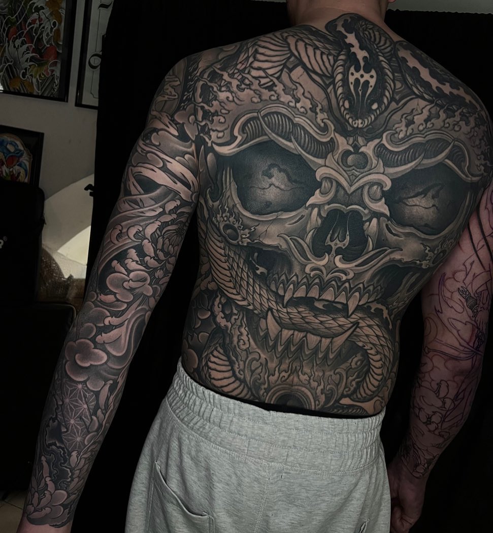 Large ornate skull with decorative baroque flourishes and a serpent, covering the back and chest in black and grey shading.