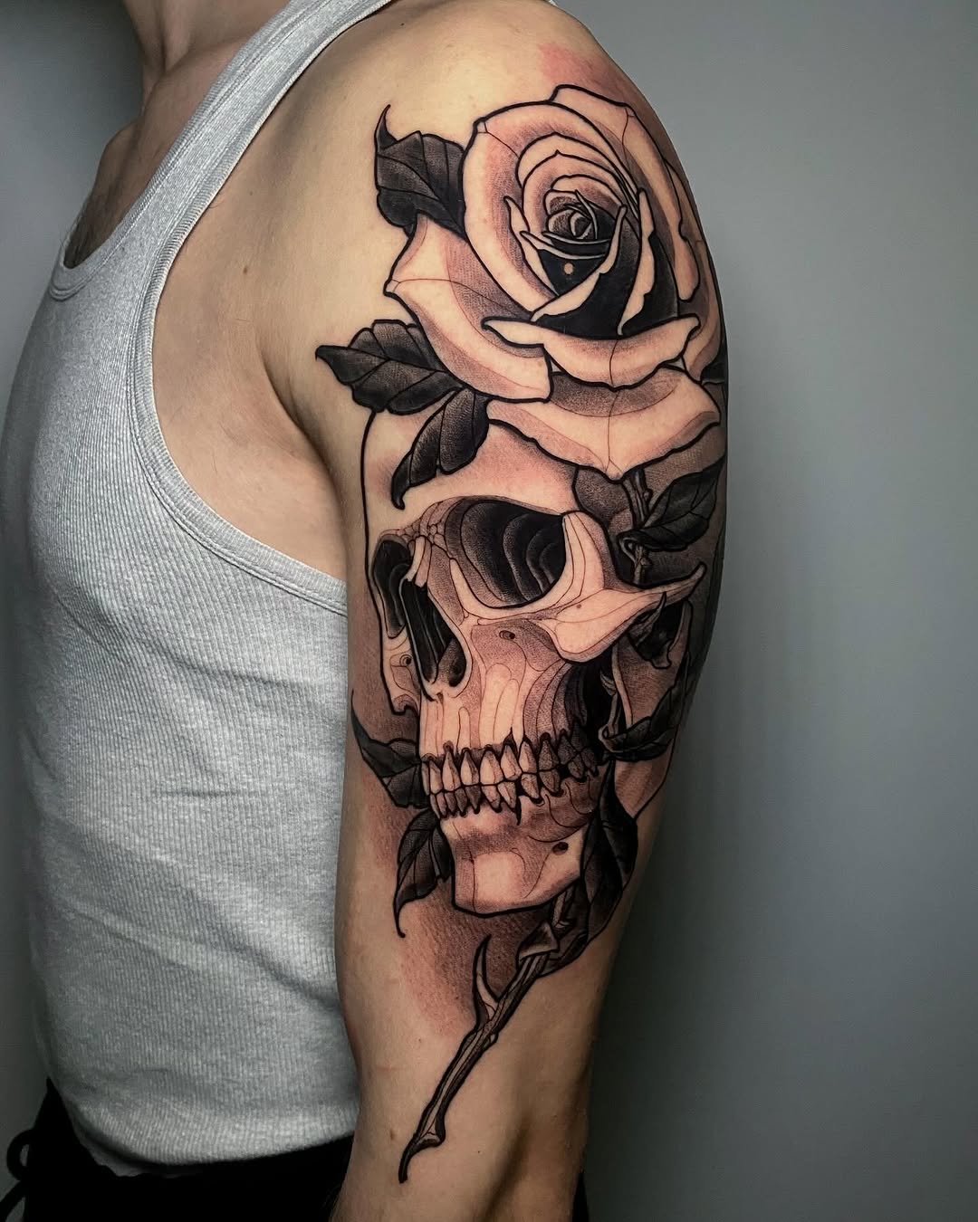 Skull with rose emerging from top on upper arm, rendered in black and grey with smooth shading and realistic detail.