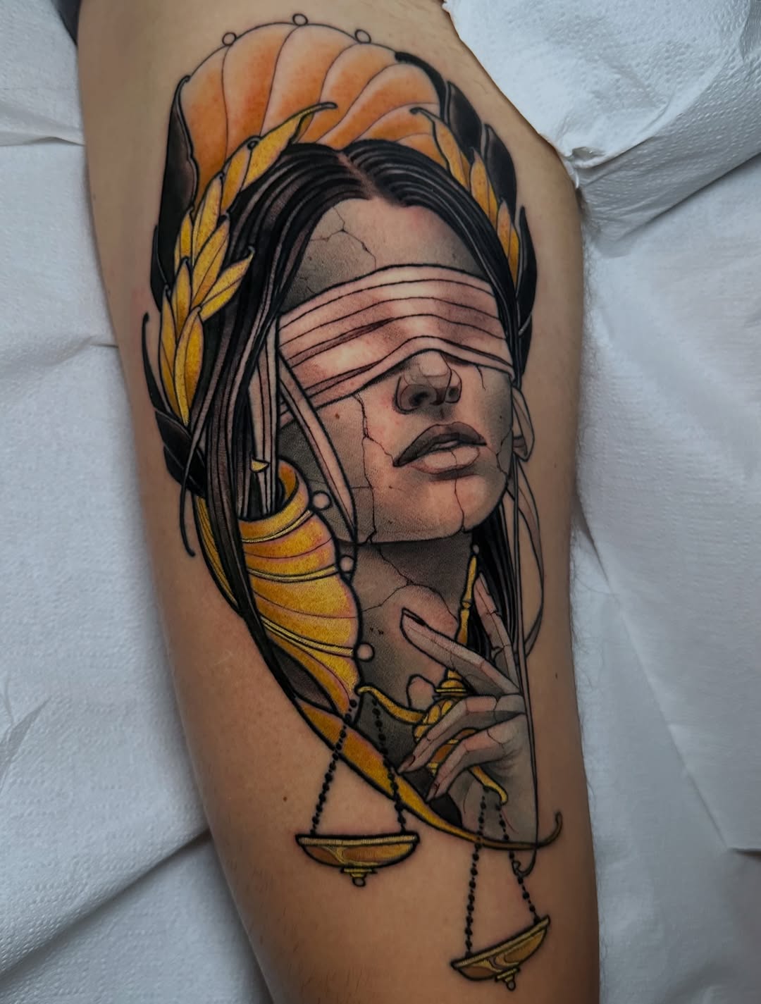 Blindfolded female portrait with flowing black hair and golden wings on upper arm, rendered in black and grey with soft shading.