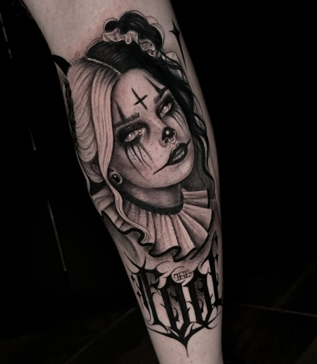 Black and grey portrait tattoo of a woman with dramatic makeup, X marks on forehead, hair in bun with scrunchie, wearing ruffled collar, on forearm.