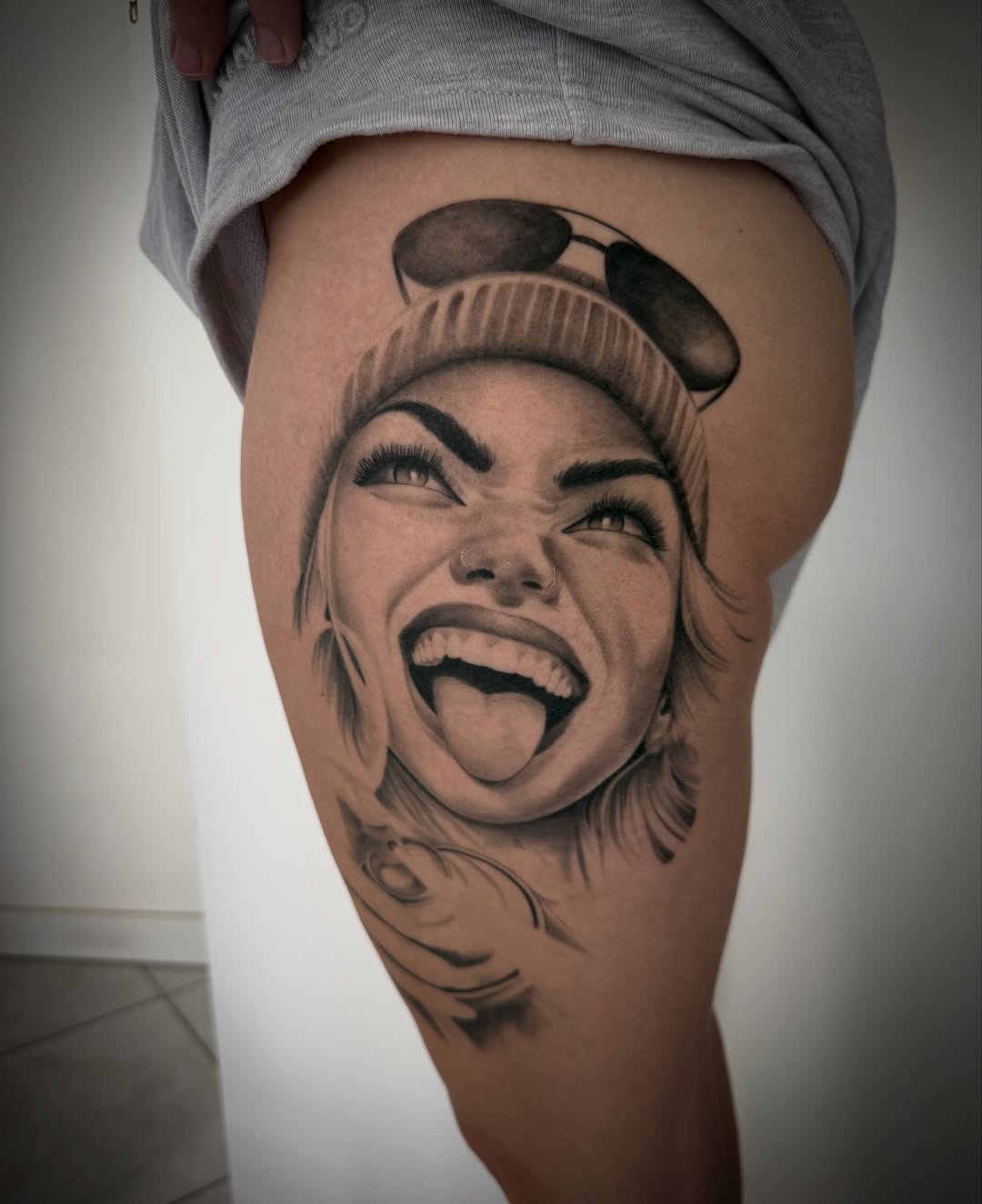 Black and grey portrait tattoo of a smiling woman wearing a beanie on the thigh, featuring detailed shading and soft gradients.