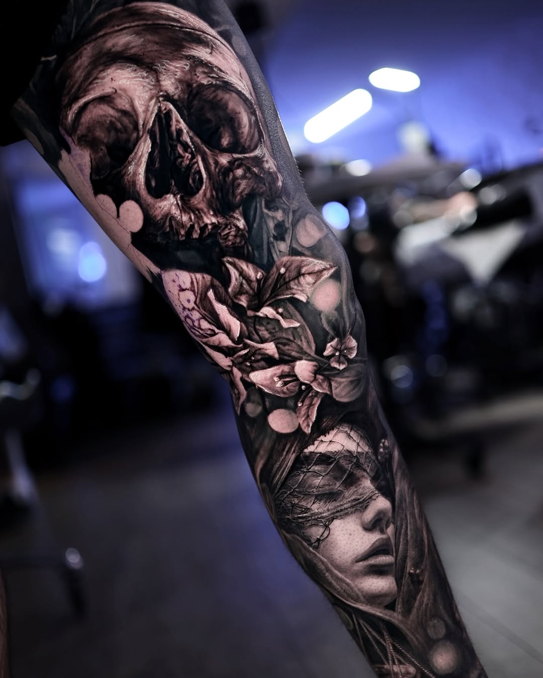 Black and grey sleeve tattoo featuring a skull with flowers and a portrait face, with detailed shading and realistic imagery on the arm.