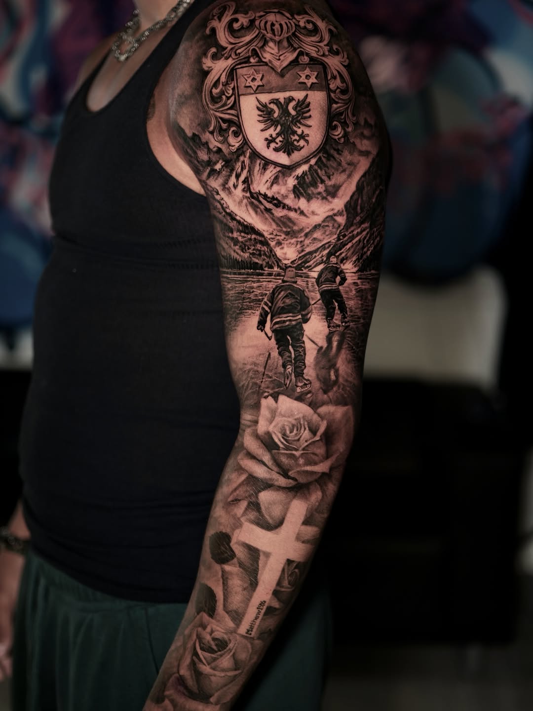 Full sleeve tattoo featuring a heraldic shield with eagle, mountainous landscape, two hikers, roses, and a cross in black and grey.