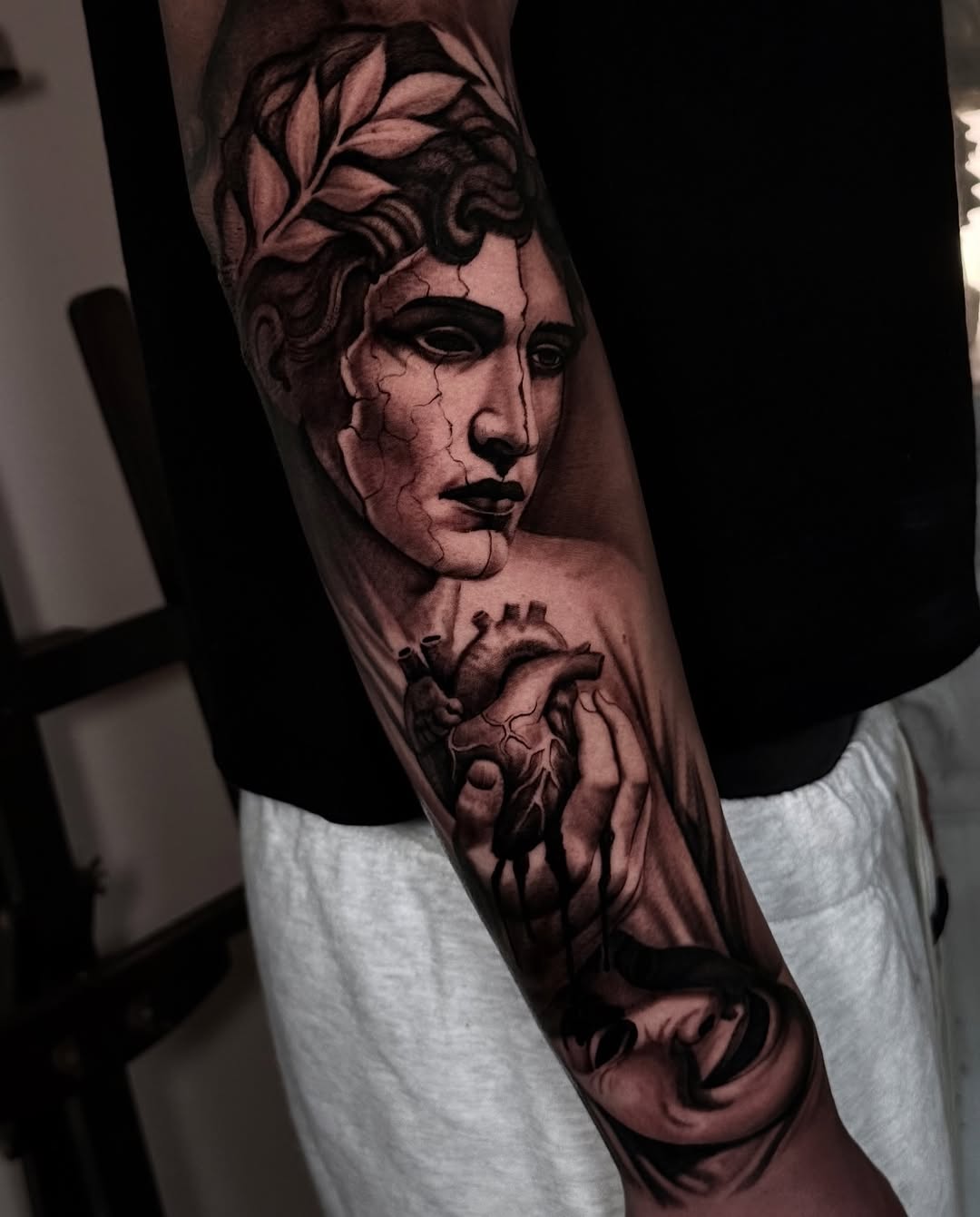 Classical male bust with laurel wreath and anatomical heart held in hands, rendered in black and grey on upper arm.