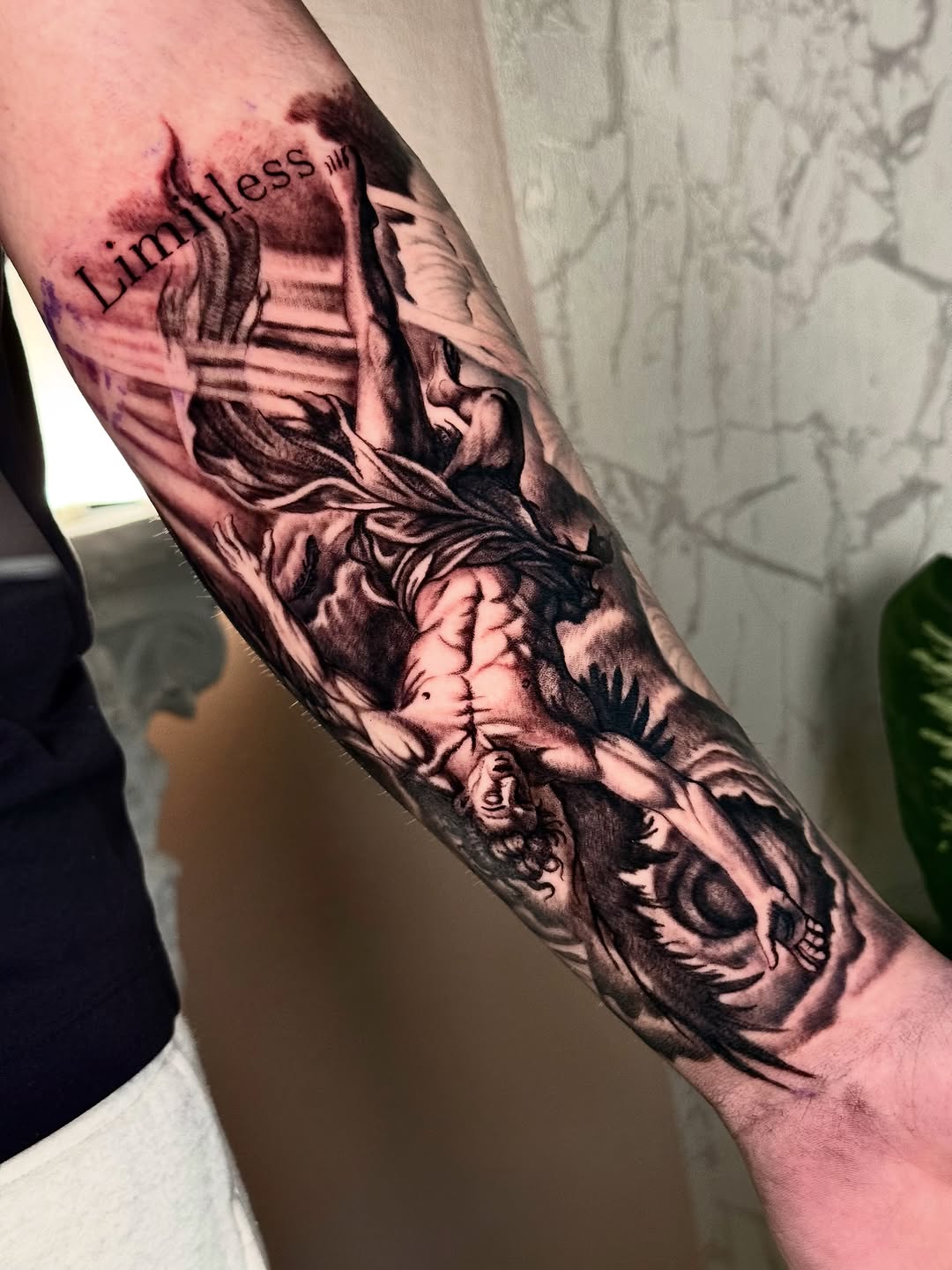 Black and grey realism tattoo on forearm featuring classical cherub angel figures with the word Limitless above, showing smooth shading and detail work.