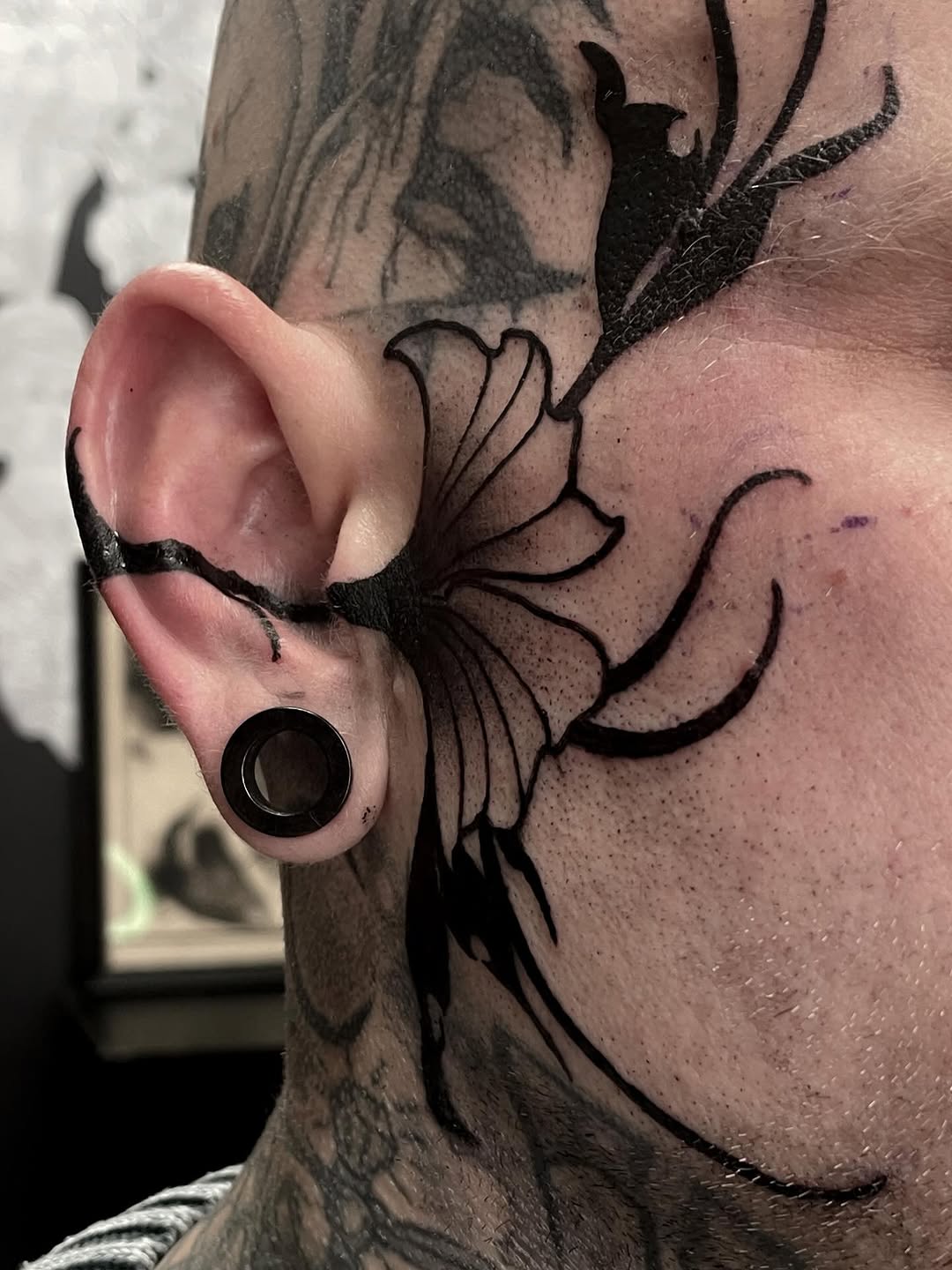 Black and grey floral tattoo on neck featuring detailed flower with flowing petals and leaves, with ear gauge visible.
