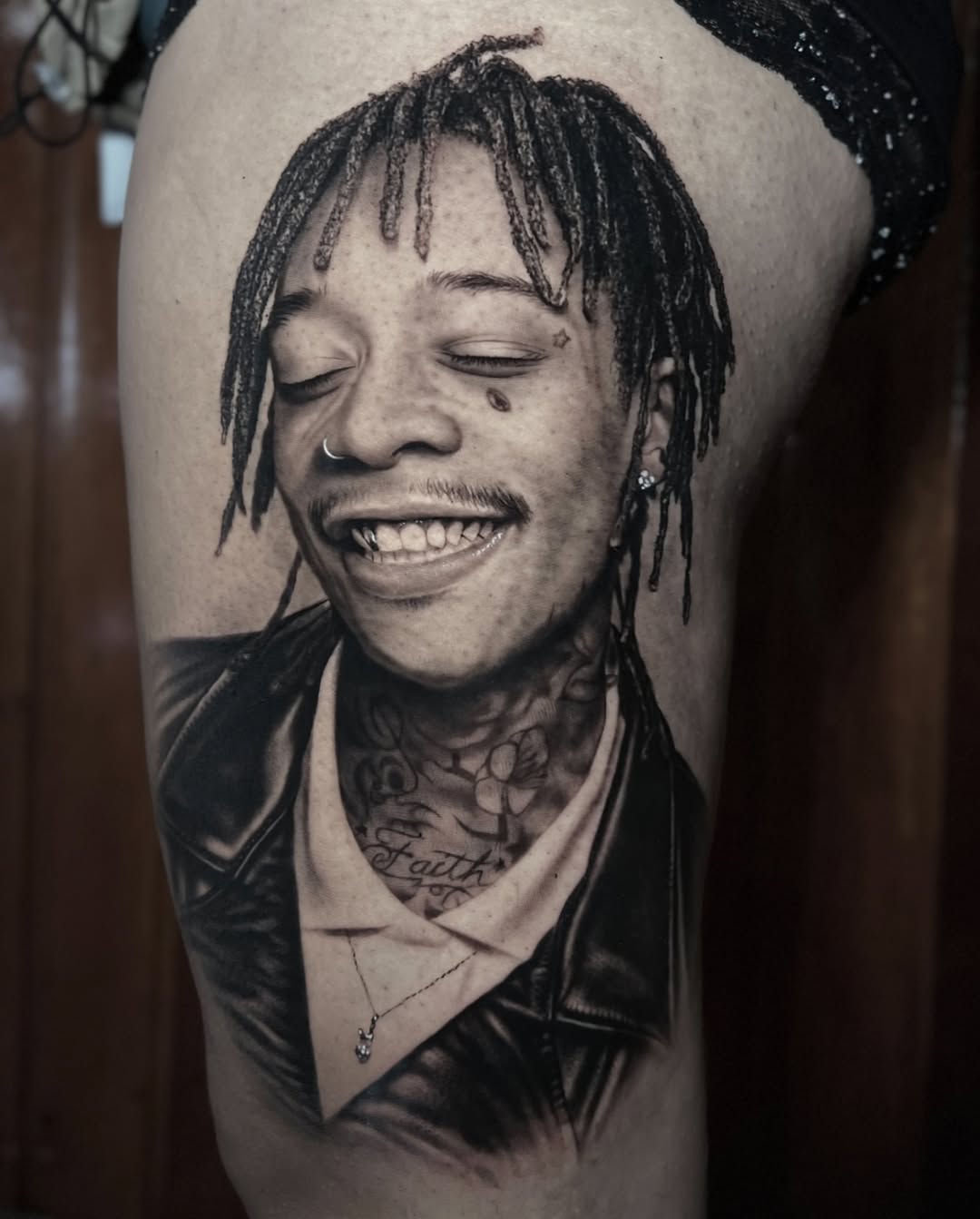 Black and grey portrait tattoo of a smiling man with braided hair and neck tattoos on upper arm, expertly shaded with soft gradients.
