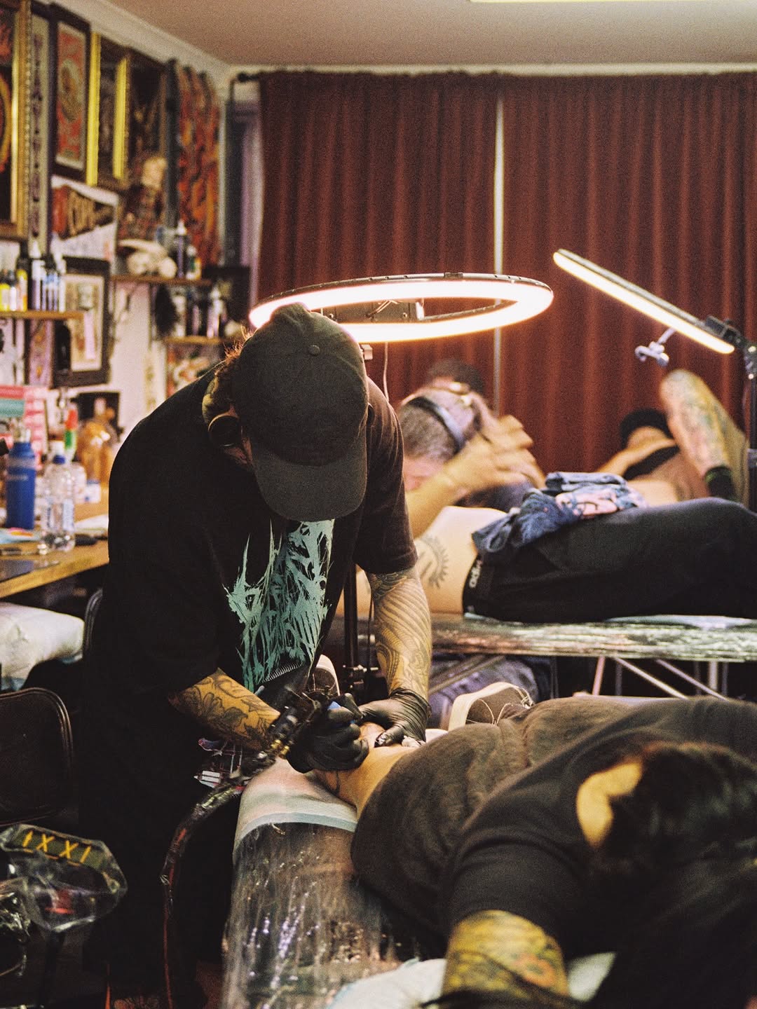 Tattoo artist working on client's leg with black and grey shading under ring light in studio.