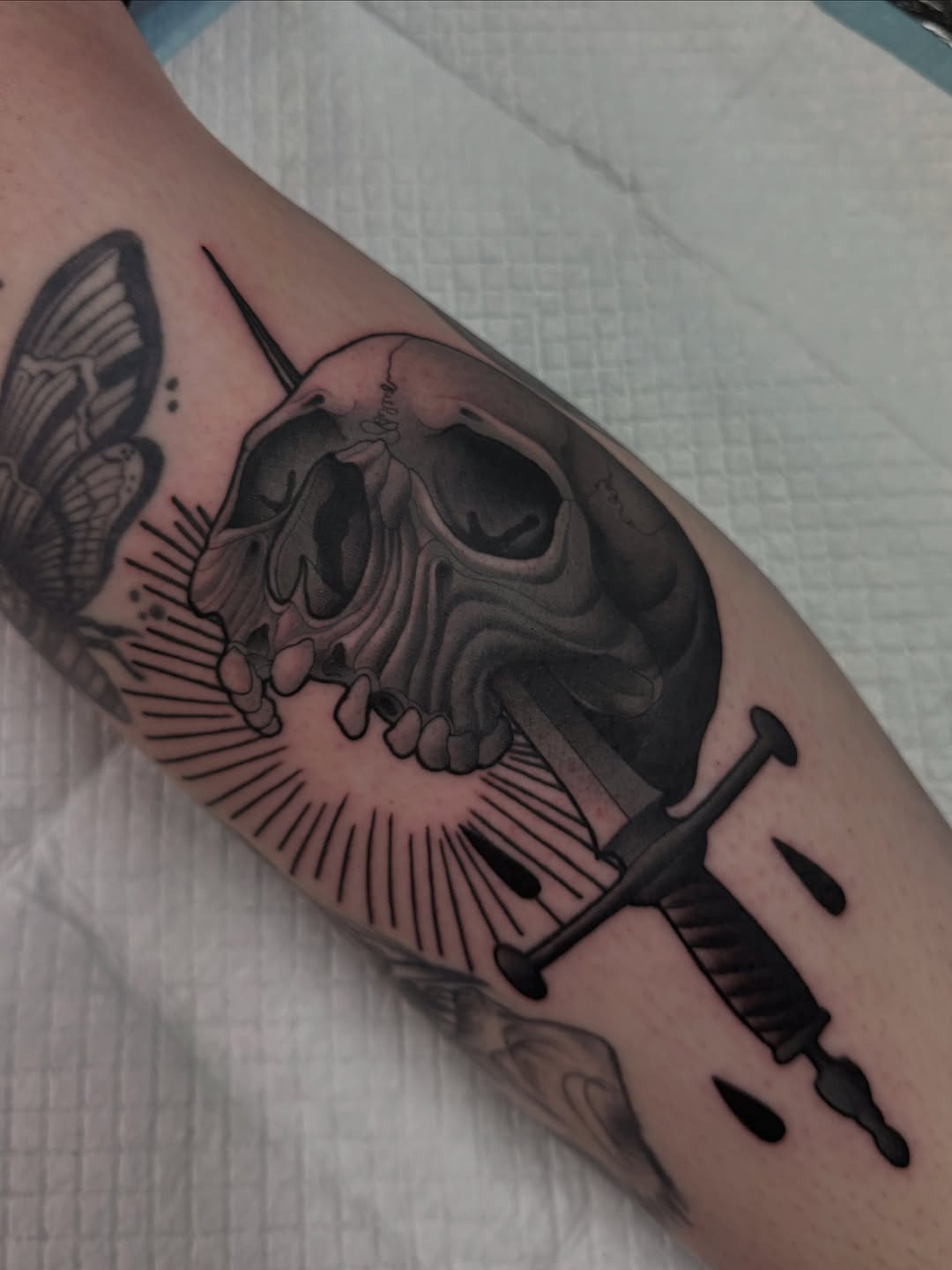 Skull with horn and wing details, shaded in black and grey, on upper arm with syringe below.